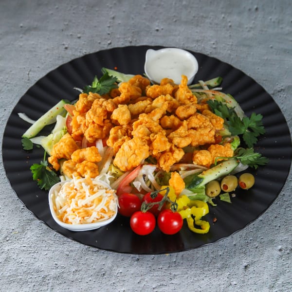 US Fried Chicken salad with crispy chicken pieces, fresh vegetables, cheese, and dressing on a black plate.