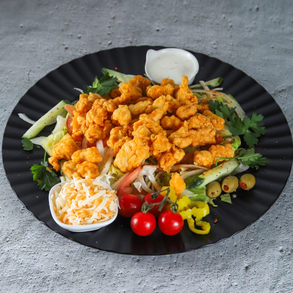 US Fried Chicken salad with crispy chicken pieces, fresh vegetables, cheese, and dressing on a black plate.