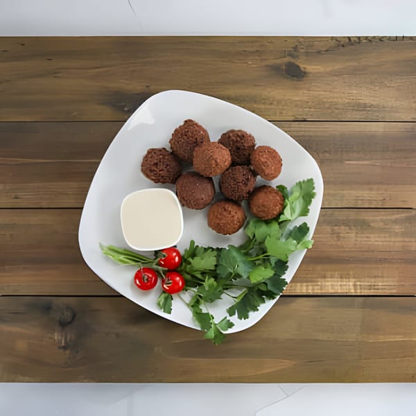 A white plate of falafel, a dipping sauce, cherry tomatoes, and parsley on a wooden table.