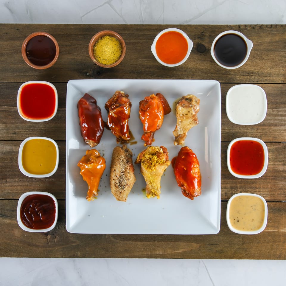 Eight US Fried Chicken wings with various sauces and seasonings, surrounded by small bowls of dipping sauces.