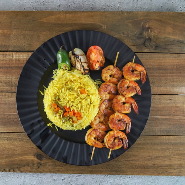 Grilled shrimp skewers, yellow rice, grilled vegetables, and a jalapeño on a black plate.