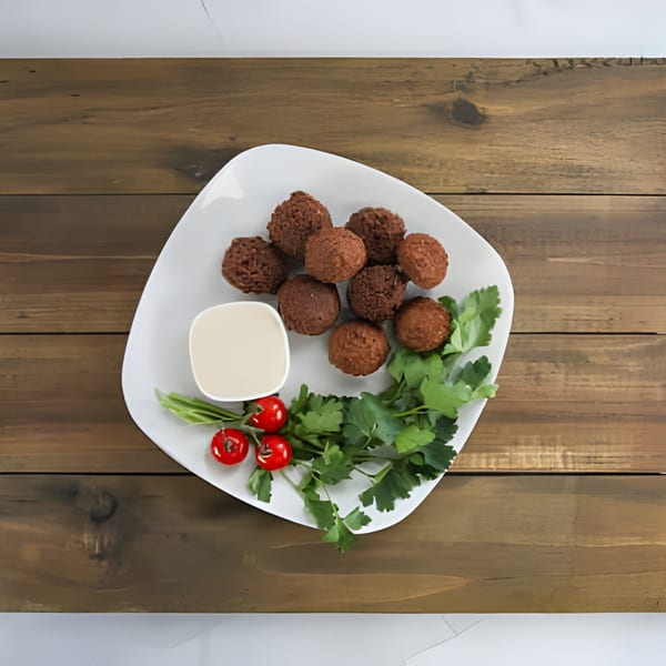 Plate of falafel, a dipping sauce, cherry tomatoes, and parsley on a wooden table.